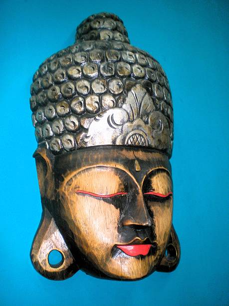 A wooden sculpture of Buddha's head against a bright blue background. The carving features a calm expression with closed eyes highlighted in red and defined lips. The intricately detailed hair is adorned with round patterns, while the forehead displays a lotus-shaped ornament. Large ear lobes are a characteristic feature, symbolizing wisdom and enlightenment. This type of sculpture is often used for spiritual or decorative purposes, reflecting traditional artistic styles in Buddhist culture.