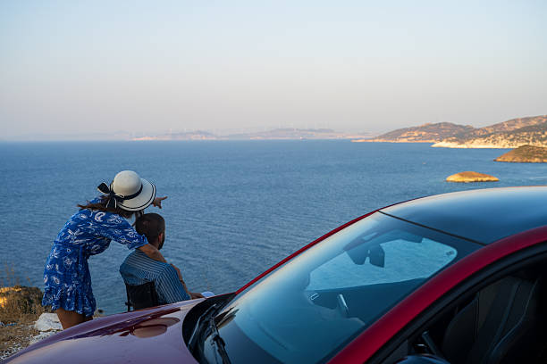 A couple enjoying the freedom of their car rental as they admire the sweeping coastal view. She leans forward with excitement, pointing towards the horizon, while he takes in the breathtaking scenery. The vibrant blue sea and warm light capture the essence of exploration and shared journeys.