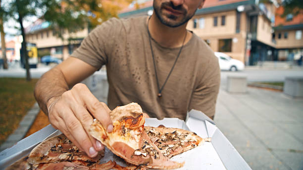 Male hand grabbing a slice from an open pizza box on a city street. The scene features a casual outdoor setting with urban background, capturing the essence of enjoying a meal in a public space.