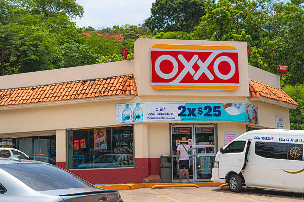 A daylight shot of a popular OXXO convenience store, recognizable by its iconic red and yellow logo. The building stands in a typical urban setting, with a person entering the store and a white van parked nearby. Palm trees and a distant hillside complete the scene, suggesting a warm, tropical climate. The image captures a familiar scene of everyday life and commerce, making it ideal for content related to travel, retail, daily life, or the culture of Mexico.