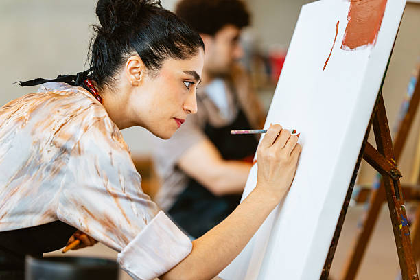 A young adult Middle Eastern female artist intensely focuses on painting a canvas during an art class. She's wearing a patterned shirt and black apron, surrounded by creative tools and attentive classmates.