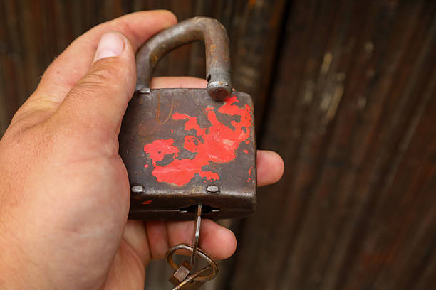​A person's hand holds an old rusty padlock with a key, symbolizing security and the passage of time. The rough metal texture and chipped red paint create a sense of history and reliability.