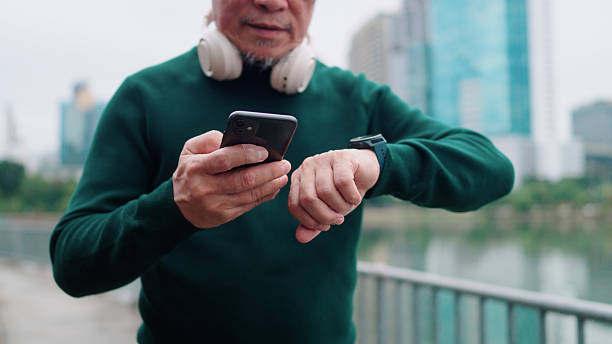 Asian elderly man connecting his smartwatch to his smartphone before starting his exercise in the park