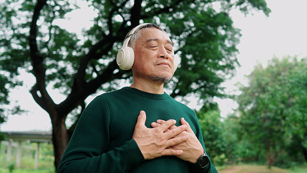 Elderly Asian man meditating under a tree with wireless headphones, calm and refreshed