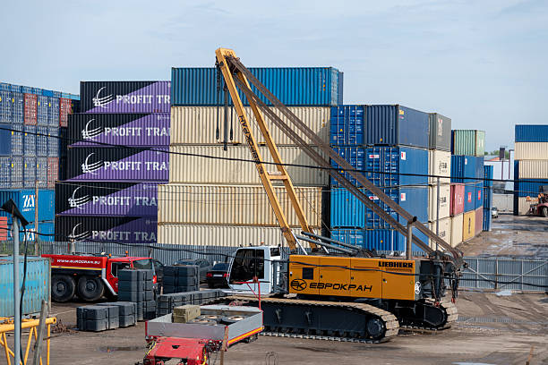 Russia, Moscow - June 09, 2025: Heavy machinery operates in a shipping yard surrounded by stacked containers. A crane carefully lifts cargo, showcasing industrial activity and logistics efficiency during daylight.