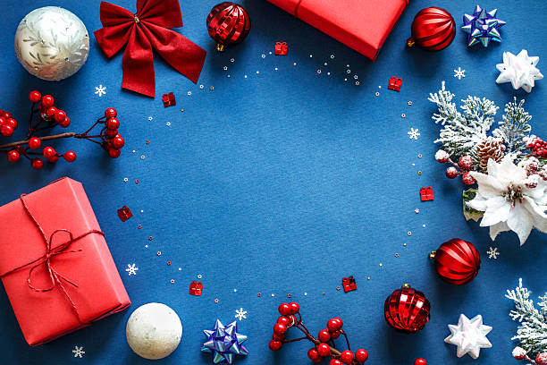 istock Festive Christmas frame with red gift boxes, snowflakes, berries, frosted pine branches, trinkets, and white and red ornaments on blue background with copy space in the middle 2235187196