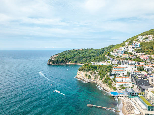 Aerial view of a coastal town with boats cruising in blue water near cliffs. Buildings densely cover the hillside overlooking the sea. Scenic oceanfront and small beach area visible.