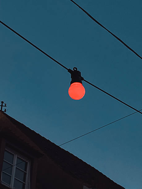 istock Red light bulb hanging from wire against a twilight sky illuminating urban atmosphere 2235207493