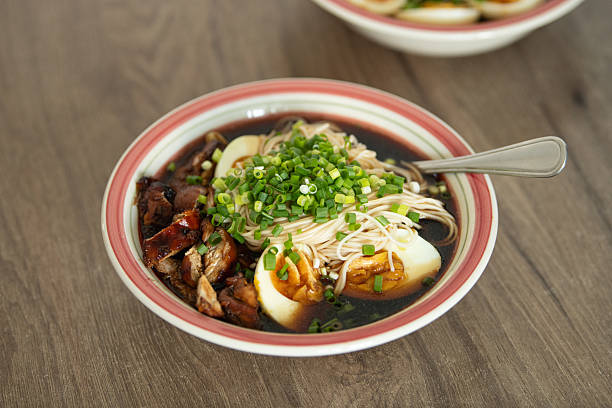 A flavorful bowl of noodles features tender chicken, sliced boiled eggs, and fresh green onions in a savory broth. Served in a warm kitchen, it's a perfect comfort meal
