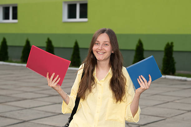 istock A joyful student stands in front of a green building, displaying two vibrant notebooks, one red and one blue. The evening light reflects her excitement for learning 2235218185