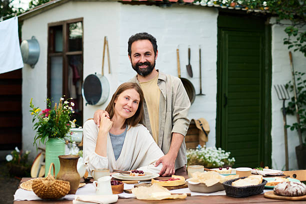 Caucasian middle aged man standing behind Caucasian middle aged woman sitting at outdoor table, both smiling and preparing food together, rustic shed and garden tools in background