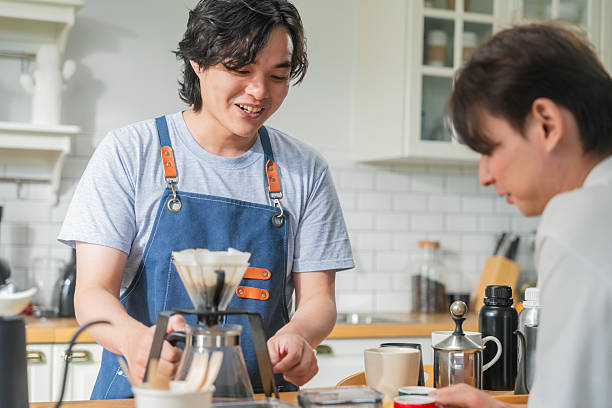 istock Asian mature man enjoys morning coffee at cafe using drip coffee bag in beige cup on white wood table, while young adult friends relax making specialty coffee as hobby activity at home 2235227172