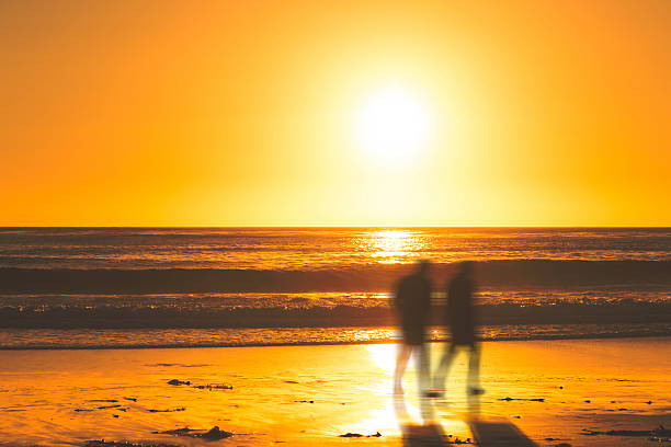 Blurry silhouettes of two people walking on Carmel Beach, California, as the sun sets over the Pacific Ocean. The intense golden glow of the sky and reflections on the wet sand create a dreamlike, abstract atmosphere. A soft motion blur adds a sense of peaceful movement and emotion.