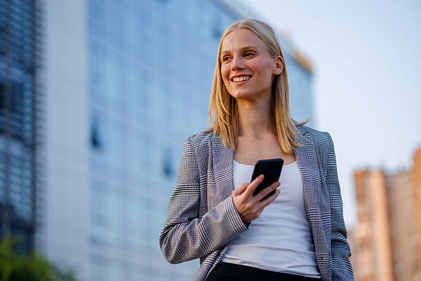istock Professional female worker checking smartphone, standing amid sleek corporate skyscrapers during sunny workday 2235239116