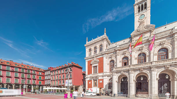 Panorama showing the town hall facade with flags in Plaza Mayor timelapse, Valladolid. Blue sky and historic architecture in the heart of the city. Pedestrians pass by in this central square.