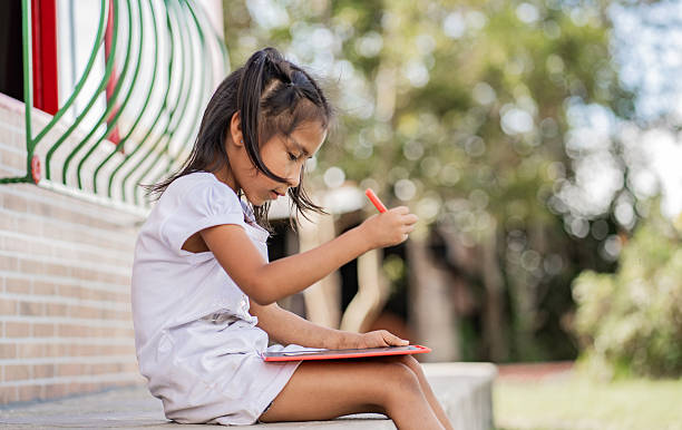 Photograph of a little girl sitting outside, dressed in white, drawing with a red marker in a notebook. Ideal image for concepts of creativity, childhood, education, learning, and child development in a natural and peaceful environment.
