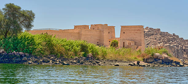 A historic temple complex along the Nile River in Egypt, framed by lush vegetation, rocky terrain, and clear blue skies, showcasing the region's cultural heritage and scenic natural beauty.