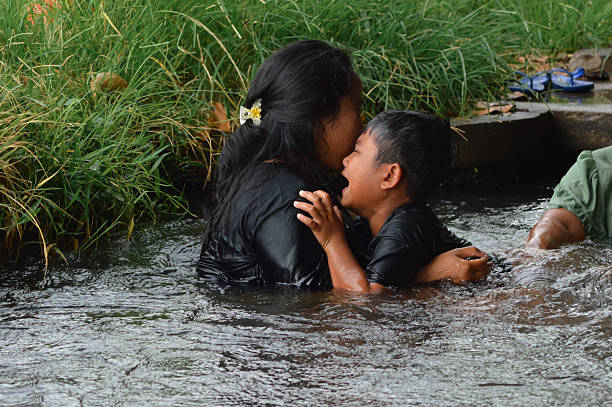 The boy clings to his mother in tears while standing in the stream, a tender countryside family moment showing comfort and emotion in the water.