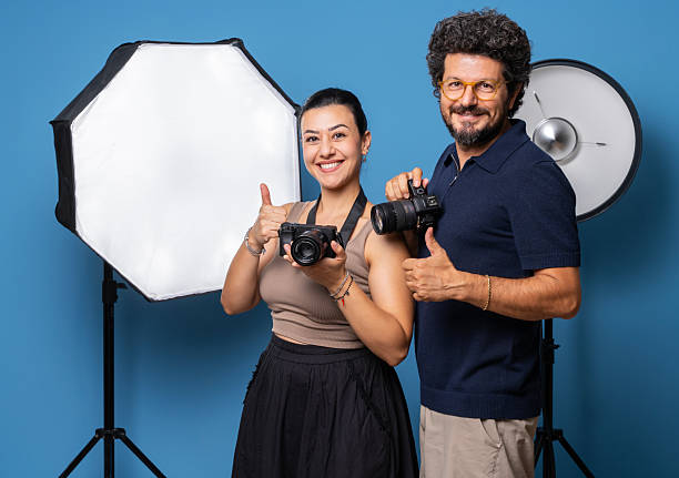 Happy male and female photographers posing with cameras in a photo studio, smiling and showing thumbs up.