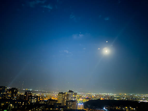 The city is lit up at night with a full moon shining brightly in the sky. The moon is positioned in the middle of the sky, casting a soft glow over the city below