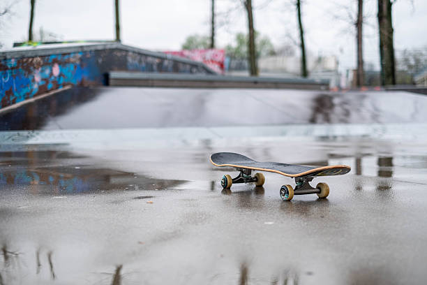 Skateboard with trucks and wheels on wet pavement reflecting ramps and graffiti in skatepark after the rain