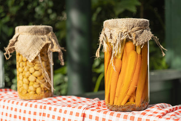 Close-up of jars with pickled carrots and chickpeas, covered with burlap lids and displayed on a red plaid tablecloth, evoking a rustic homemade harvest vibe