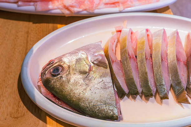 This is a close-up photograph of a raw fish, meticulously sliced and presented on a white oval plate. The head of the fish is displayed on the left, leading to a series of uniform, thick slices that reveal the fresh, pale flesh within. The image captures the natural, iridescent sheen of the fish's skin under bright lighting, highlighting its freshness and readiness for cooking, likely for a hot pot or steam dish. The shallow depth of field softly blurs another platter of food in the background, suggesting a communal dining experience.