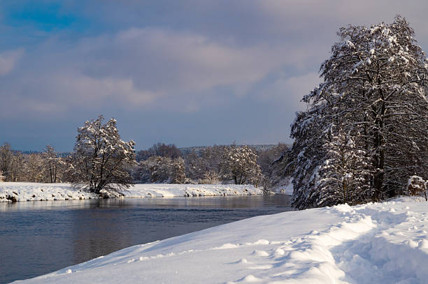Scenic winter landscape with snow-covered ground, frozen riverbank and frosty trees under a bright blue sky.