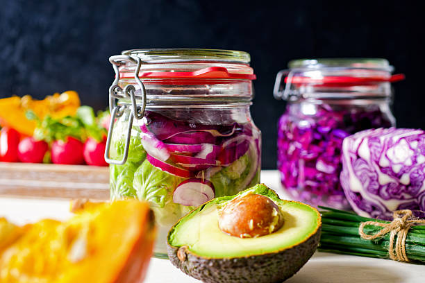 Two glass jars filled with vegetables sit on a countertop. One jar contains Brussels sprouts and red onion. The other jar contains red cabbage. Vegetables and Avocado are in front. Fermented vegetables for probiotic health. Close-up - food and drink, wellbeing