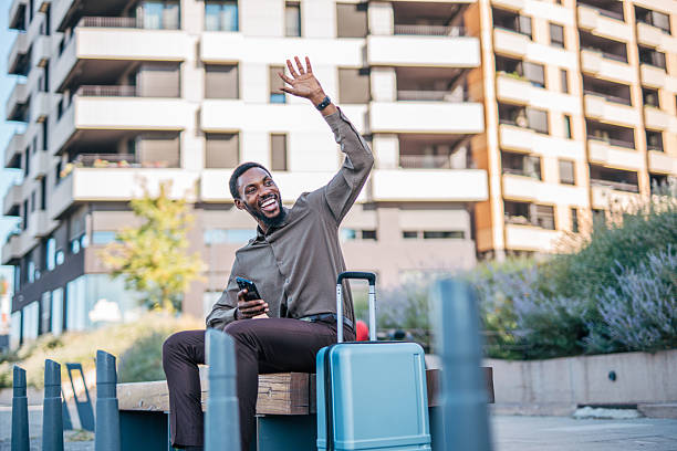 istock Smiling businessman waving goodbye while sitting on bench with suitcase 2235354385