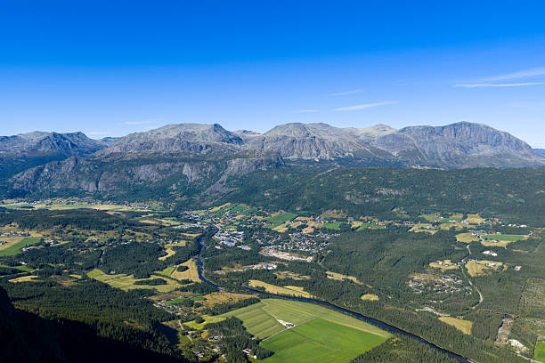 A wide aerial photograph of an idyllic Norwegian valley. A dramatic, rugged mountain range forms a powerful backdrop, while a small town with a winding river and patches of green farmland and dense forests fills the foreground. Under a vast, clear blue sky, this image beautifully captures the harmony between human settlement and the raw, magnificent power of nature. This shot is perfect for travel, tourism, and real estate-themed projects.