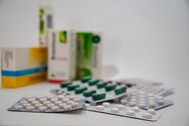 Blister packs of tablets and capsules displayed in the foreground with blurred medicine boxes in the background on white surface.