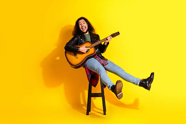 istock Attractive stylish man playing guitar wearing a leather jacket, seated against a bright yellow background, full of energy 2235367707