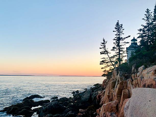 A lighthouse stands perched on rugged rocky cliffs, overlooking a calm ocean at sunset. Silhouetted pine trees and a pastel-colored sky create a peaceful maritime landscape, ideal for travel and nature themes.