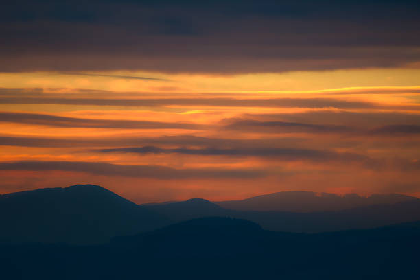A minimalist, telephoto shot of a sunset or sunrise sky featuring dramatic, layered clouds painted in rich tones of dark orange, gold, and deep purple-blue. The lower third of the frame shows a dark silhouette of rolling hills and mountains fading into the distance, with the stratified sky dominating the composition and creating a sense of moody atmosphere and warmth.