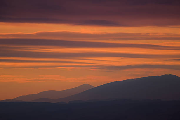 A horizontal, telephoto image capturing the warm colors of a sunrise or sunset over a layered mountain range. The sky is filled with delicate, horizontal streaks of bright orange, gold, and soft purple clouds. The lower portion of the image features sharp, dark silhouettes of rolling mountains, emphasizing the contrast between the warm, layered atmosphere above and the cool, shadowed landscape below.