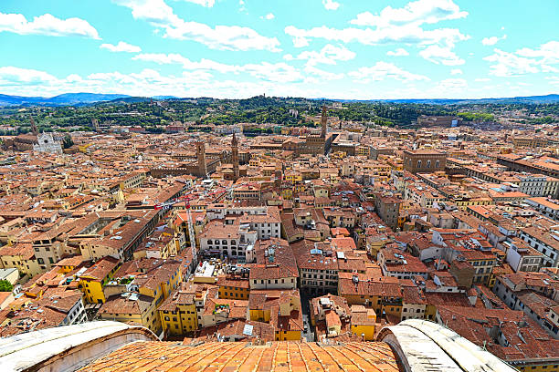 A wide, elevated perspective capturing the dense, sprawling historic center of Florence, Italy. The view looks down over the tightly clustered terracotta-tiled rooftops and narrow streets, extending to the green hills and surrounding Tuscan landscape in the distance. The bright blue sky is dotted with soft white clouds, and the composition is framed in the foreground by the edge of a curved, tiled dome (likely the Duomo or Campanile).