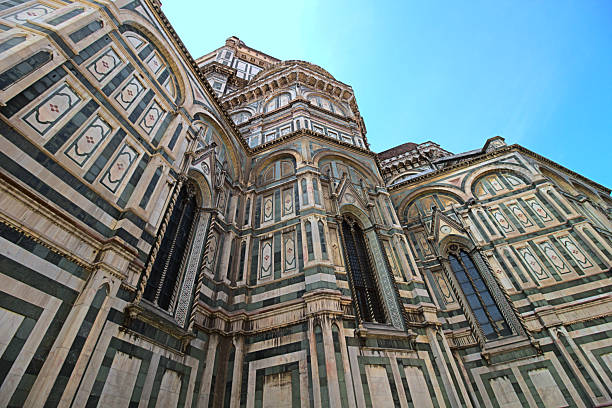 A dramatically low-angle, wide-view shot of the patterned exterior of the Florence Duomo (Cattedrale di Santa Maria del Fiore) in Italy. The massive Gothic structure features intricate geometric designs in green, white, and pink marble, rising steeply toward a bright blue sky. The ornate windows and the distinct striped facade dominate the frame, emphasizing the monumental scale and Renaissance architecture of the bell tower (Giotto's Campanile) section.