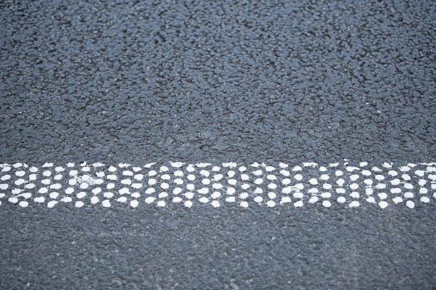A white dotted line is painted on a black asphalt road surface, clearly marking the lanes in a busy urban environment during daylight. Vehicles can be seen in the background.