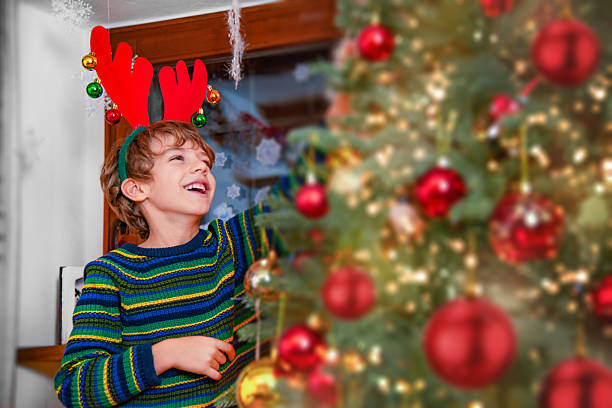 Happy boy wearing festive reindeer antlers while decorating a Christmas tree with red ornaments and lights. Warm holiday atmosphere at home with laughter and celebration.