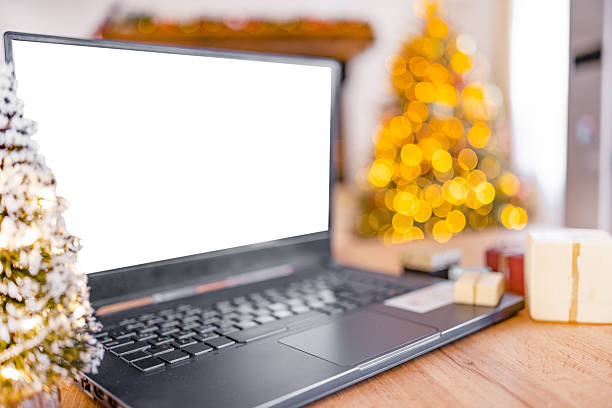 Modern laptop with blank white screen on wooden table, decorated Christmas tree with glowing lights in the background. Concept of online shopping, e-commerce, smart working, digital greetings, and holiday season technology.