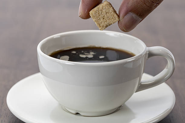 Hand holding organic sugar cube above hot black coffee in white ceramic cup on saucer, wooden table background, morning beverage concept, cozy and inviting atmosphere