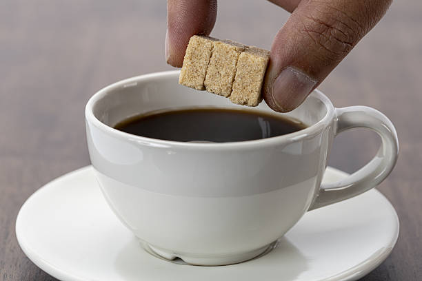 Hand adding three organic sugar cubes into cup of black coffee, creating cozy and inviting atmosphere on wooden table with white ceramic cup and saucer