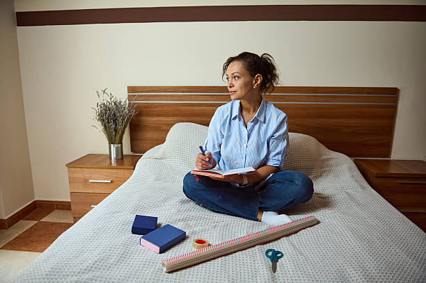 istock Woman Sitting on Bed Writing Notes Surrounded by Creative Supplies 2235396269