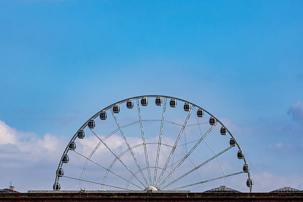A large, white Ferris wheel with dark gondolas is set against a bright blue sky with sparse clouds, rising above a brick structure  in Liverpool, UK.