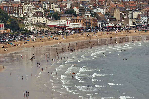 A bustling beach scene with many people, buildings, a Ferris wheel, and the ocean with waves  in Scarborough, UK.