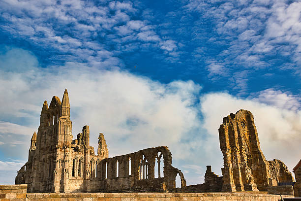 Ruined stone abbey against a dramatic blue sky with wispy white clouds. Sunlight illuminates the weathered architecture  in Whitby, UK.