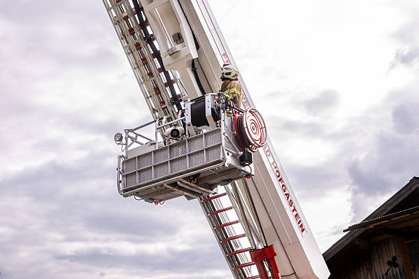 A fire truck equipped with a long ladder is parked near a residential building. Firefighters in full gear are working near the vehicle, preparing for an operation. The scene is set in a residential area with a sidewalk, grass, and parked cars.