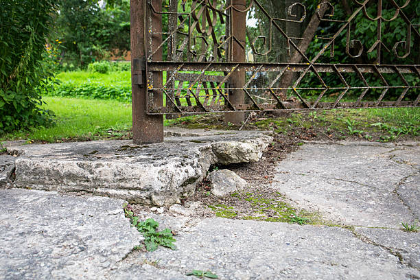 A weathered metal gate attached to a rusty pole standing on broken and crumbling concrete base in a garden entrance.