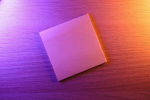 A top-down view of a blank, square piece of paper resting on a wooden desk. The scene is lit with dramatic, contrasting light, casting a vibrant purple glow on the left and a warm orange hue on the right. This unique lighting creates an artistic and moody atmosphere. The clean, blank surface is ideal as a creative background or a mockup for text, logos, or product presentations.